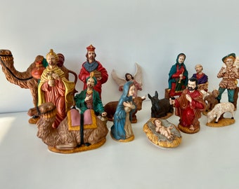 May include: A nativity scene set with 13 ceramic figurines, including the three wise men, Mary, Joseph, baby Jesus, an angel, a donkey, a camel, and sheep. The figurines are painted in bright colors and have detailed features.