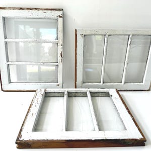May include: Three rectangular wooden window frames with distressed white paint finish. Each frame features three glass panes separated by thin muntins. The wood shows signs of wear with exposed brown grain along the edges and corners of the frames.