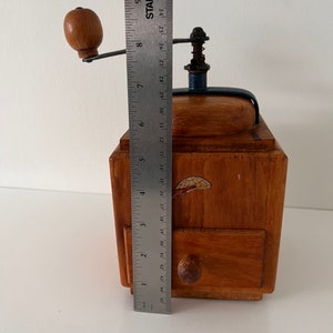Puede incluir: Un molinillo de caf&eacute; de madera con una manivela de metal y un peque&ntilde;o caj&oacute;n con un pomo de madera. Una regla de metal est&aacute; colocada al frente, mostrando que la altura del molinillo es de aproximadamente 23 cm desde la base hasta la manivela.