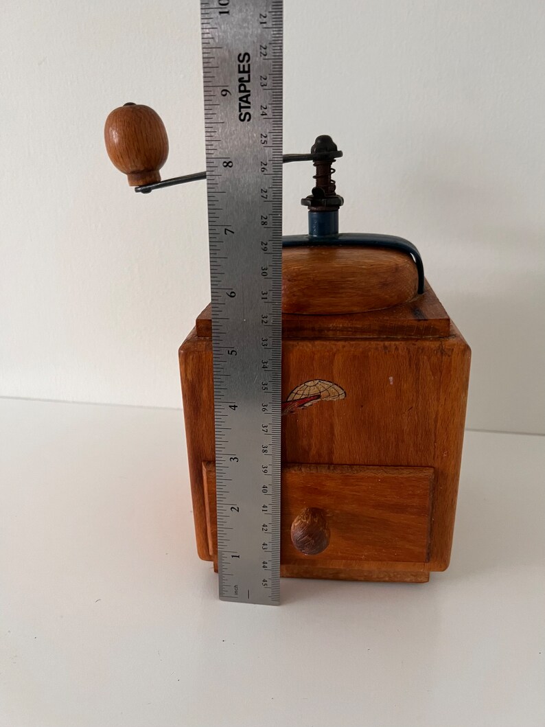 Puede incluir: Un molinillo de caf&eacute; de madera con una manivela de metal y un peque&ntilde;o caj&oacute;n con un pomo de madera. Una regla de metal est&aacute; colocada al frente, mostrando que la altura del molinillo es de aproximadamente 23 cm desde la base hasta la manivela.