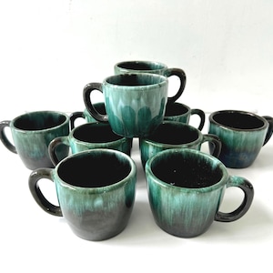 May include: A set of ten ceramic mugs with a distinctive glaze. The mugs are a mix of teal and dark grey, with a glossy finish. Each mug has a rounded body, a dark handle, and a dark interior. The mugs are stacked and arranged on a white surface.