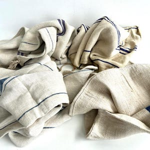 May include: Several beige linen towels with dark blue stripes are arranged on a white surface. The towels are folded and draped, showcasing their texture and design. The towels have a vintage, rustic aesthetic.