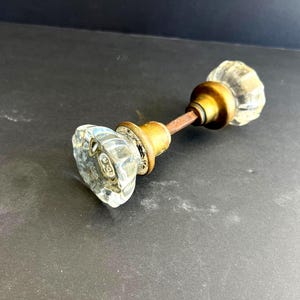 May include: A clear glass door knob with a faceted design and brass hardware. The knob has a vintage aesthetic and is attached to a metal rod. The door knob is a classic design.