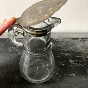 May include: A clear glass pitcher with a metal lid. The lid is hinged and has a handle. The pitcher has a textured surface and a spout.