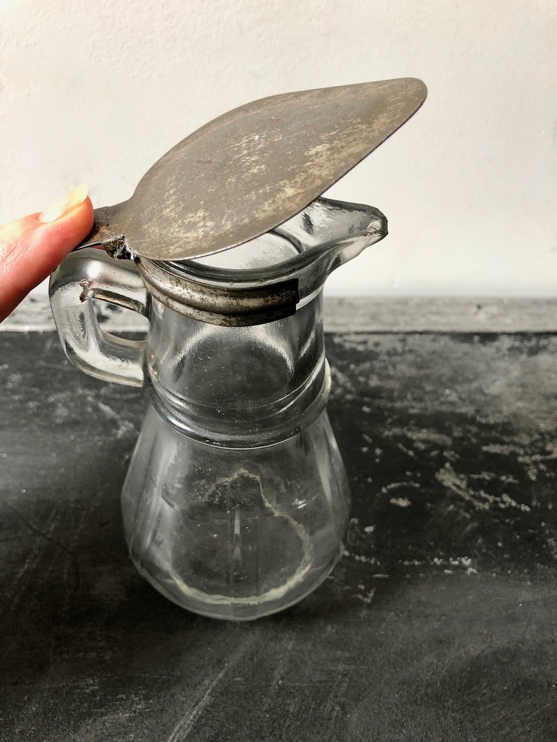 May include: A clear glass pitcher with a metal lid. The lid is hinged and has a handle. The pitcher has a textured surface and a spout.