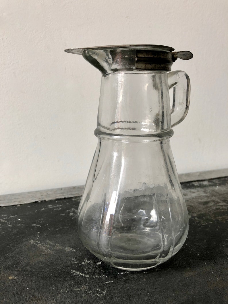 May include: A clear glass pitcher with a silver lid and a handle. The pitcher has a textured, ribbed design.