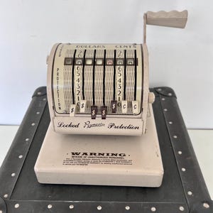 May include: A vintage white cash register with a black handle and a black case. The register has a display that reads "DOLLARS" and "CENTS" and has a warning label that reads "WARNING. BEWARE OF UNAUTHORIZED PERSONNEL. Only a Paymaster Technician can service or repair the Paymaster 2 YEAR WARRANTY is void if unauthorized personnel is allowed to service your PAYMASTER."