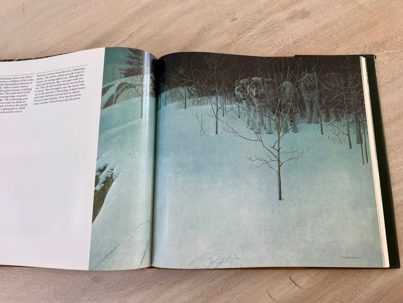 May include: A book open to a page with a painting of a snowy forest scene. The painting depicts a pack of wolves standing in the snow, with trees and bushes behind them. The text on the page reads "However, I do not see them as signifying menace. The shadows, which cast themselves over the snow, are not menacing. Although they are how and where they are, you never feel that they are on the attack. You never feel that they are a threat. They are doing what wolves do, and they are doing it in a way that is both beautiful and striking. Now the feeling of danger comes only from the viewer, and the viewer alone will make that decision."