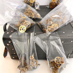 May include: Several clear plastic bags filled with small, brass-colored clock gears and components. The bags are arranged on a dark, textured surface, possibly a vintage suitcase. The gears vary in size and shape, suggesting a collection of watch or clock parts.