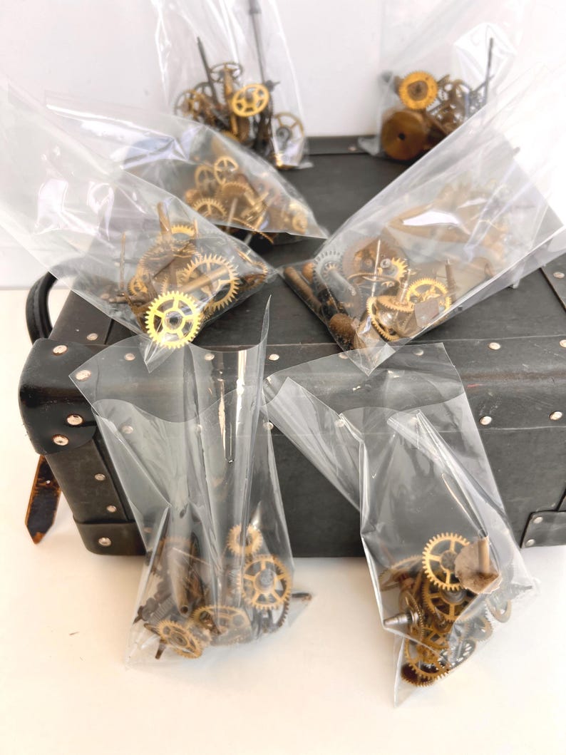 May include: Several clear plastic bags filled with small, brass-colored clock gears and components. The bags are arranged on a dark, textured surface, possibly a vintage suitcase. The gears vary in size and shape, suggesting a collection of watch or clock parts.