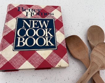May include: A red and white checkered cookbook titled "Better Homes and Gardens New Cookbook". Three wooden spoons are on a white countertop next to the book.