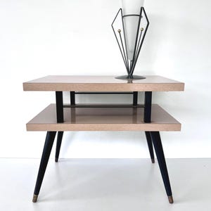 May include: A mid-century modern two-tier side table with a light wood grain finish. The table has black legs and supports, with a decorative metal accent. A black metal and glass decorative piece sits on top.