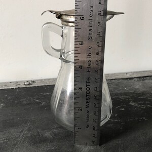 May include: A clear glass bottle with a metal lid and a handle. The bottle is about 6 inches tall and has a pear-shaped body. The bottle is sitting on a black surface next to a ruler.