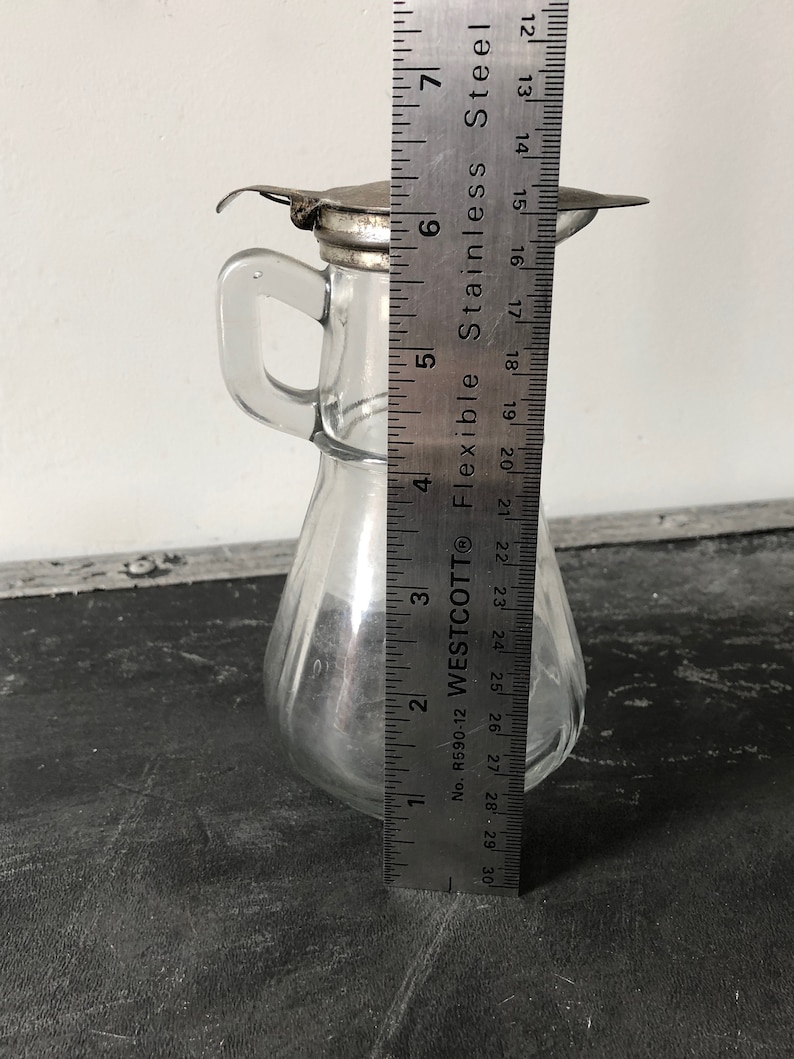 May include: A clear glass bottle with a metal lid and a handle. The bottle is about 6 inches tall and has a pear-shaped body. The bottle is sitting on a black surface next to a ruler.