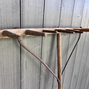 May include: A vintage wooden rake head with several tines and a long wooden handle. The rake head is made of aged wood with metal tines. The handle is attached with metal supports. The rake is designed for gardening.