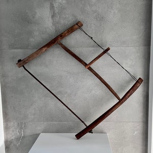 May include: A wooden saw with a metal blade, hanging on a grey wall. The saw is made of dark wood and has a simple design.