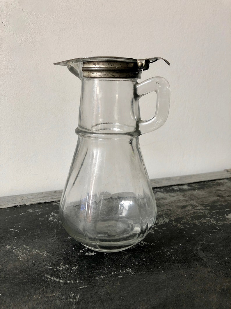 May include: A clear glass pitcher with a silver lid and a handle. The pitcher has a textured, ribbed design.
