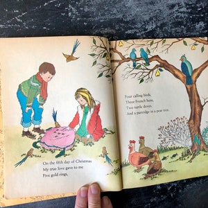 May include: An open book with colorful illustrations of a girl and boy holding a pink object with gold rings. The text on the page reads "On the fifth day of Christmas My true love gave to me Five gold rings, Four calling birds, Three French hens, Two turtle doves, And a partridge in a pear tree."