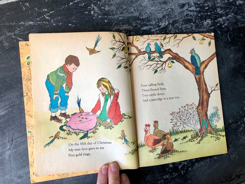 May include: An open book with colorful illustrations of a girl and boy holding a pink object with gold rings. The text on the page reads "On the fifth day of Christmas My true love gave to me Five gold rings, Four calling birds, Three French hens, Two turtle doves, And a partridge in a pear tree."