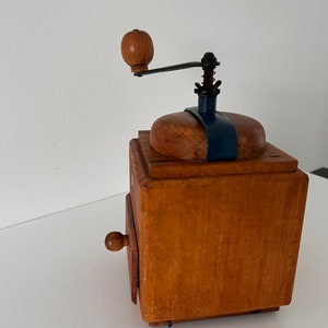 Puede incluir: Un molinillo de caf&eacute; cuadrado de madera con acabado marr&oacute;n claro, manivela de metal oscuro con pomo de madera redondo, soporte superior de metal azul y un peque&ntilde;o caj&oacute;n extra&iacute;ble con un pomo de madera redondo a juego en la parte frontal.