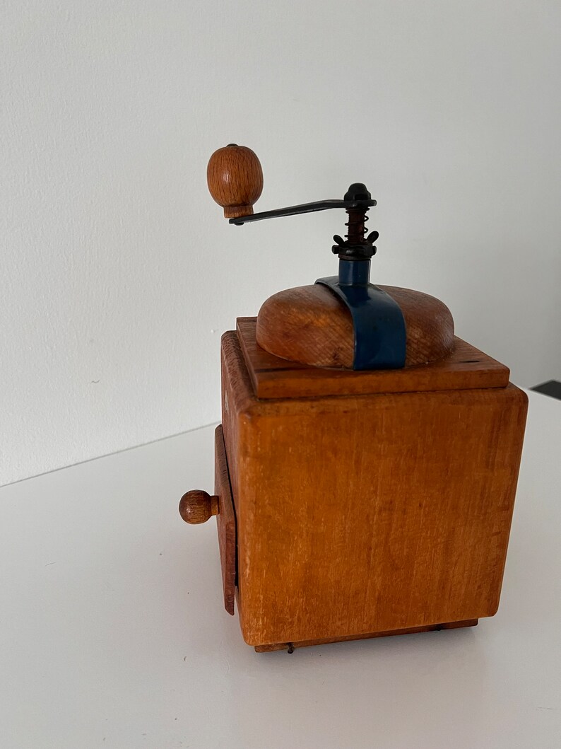 Puede incluir: Un molinillo de caf&eacute; cuadrado de madera con acabado marr&oacute;n claro, manivela de metal oscuro con pomo de madera redondo, soporte superior de metal azul y un peque&ntilde;o caj&oacute;n extra&iacute;ble con un pomo de madera redondo a juego en la parte frontal.