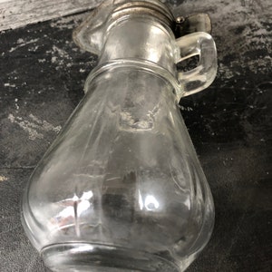 May include: A clear glass pitcher with a silver lid and handle. The pitcher has a rounded body and a textured surface.