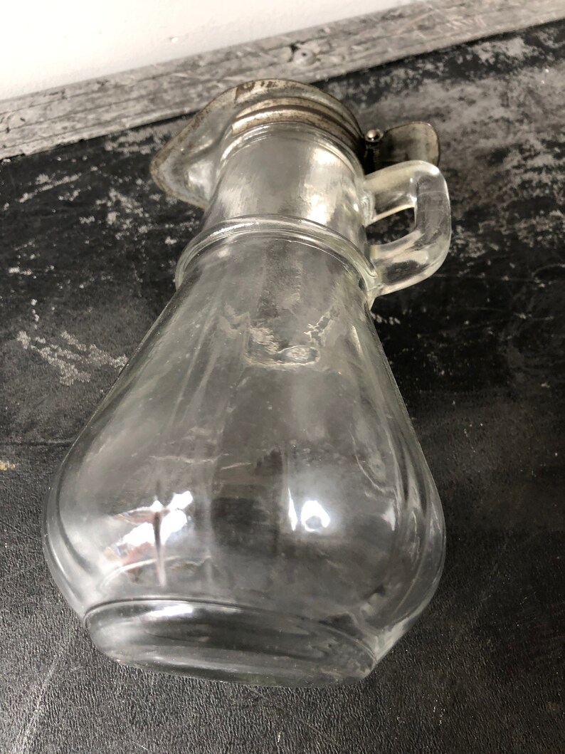 May include: A clear glass pitcher with a silver lid and handle. The pitcher has a rounded body and a textured surface.