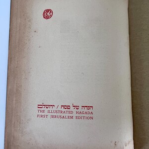 Puede incluir: Primer plano de la portada de un libro titulado "The Illustrated Haggadah, First Jerusalem Edition". La portada es de color marr&oacute;n claro con un sello rojo en la esquina superior izquierda.