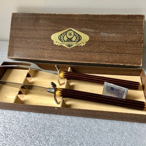 May include: A wooden box with a brown lid containing a carving knife and fork set with brown and gold handles. The box has a gold label that reads "The Connoisseur's Choice".