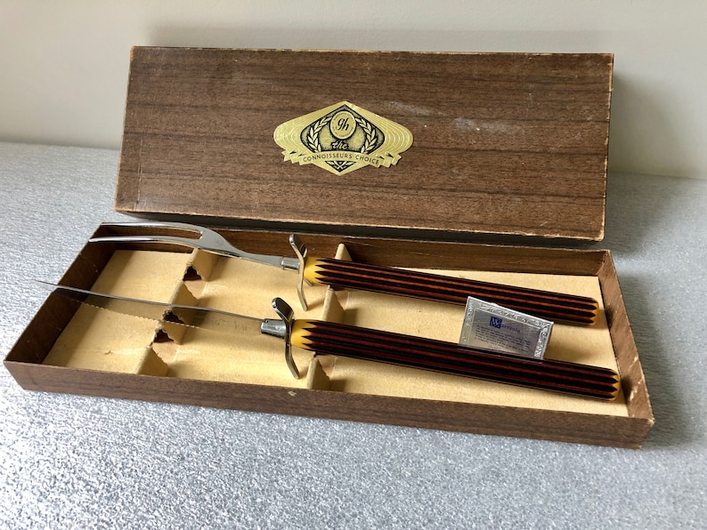 May include: A wooden box with a brown lid containing a carving knife and fork set with brown and gold handles. The box has a gold label that reads "The Connoisseur's Choice".