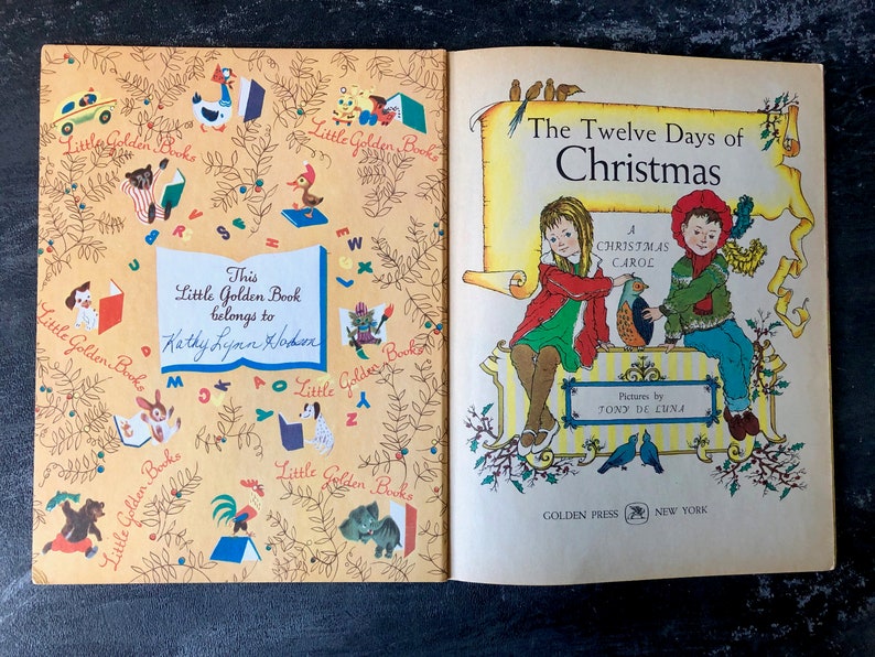 May include: An open book with a yellow cover and illustrations of animals and letters. The book is titled "The Twelve Days of Christmas" and features a Christmas carol. The book is a Little Golden Book and has a personalized inscription.