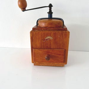 Puede incluir: Un molinillo de caf&eacute; cuadrado de madera con acabado en marr&oacute;n claro, que cuenta con una manivela de metal negro con pomo de madera, una tapa superior y un peque&ntilde;o caj&oacute;n extra&iacute;ble en la base. Un peque&ntilde;o gr&aacute;fico de un paraca&iacute;das se ve en el frente.