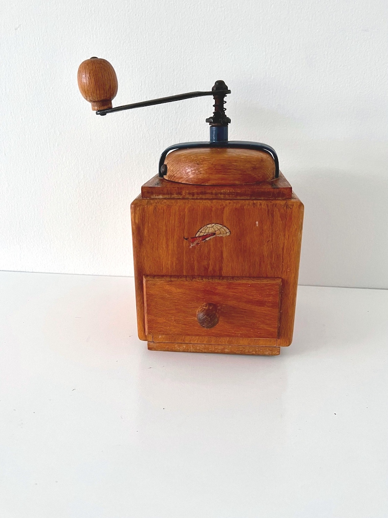 Puede incluir: Un molinillo de caf&eacute; cuadrado de madera con acabado en marr&oacute;n claro, que cuenta con una manivela de metal negro con pomo de madera, una tapa superior y un peque&ntilde;o caj&oacute;n extra&iacute;ble en la base. Un peque&ntilde;o gr&aacute;fico de un paraca&iacute;das se ve en el frente.