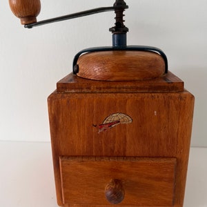 Puede incluir: Un molinillo de caf&eacute; de madera con cuerpo cuadrado, un peque&ntilde;o caj&oacute;n extra&iacute;ble con pomo redondo y una manivela de metal montada en la parte superior con un pomo de madera. El frente de la caja de madera muestra un peque&ntilde;o gr&aacute;fico desgastado de un globo.