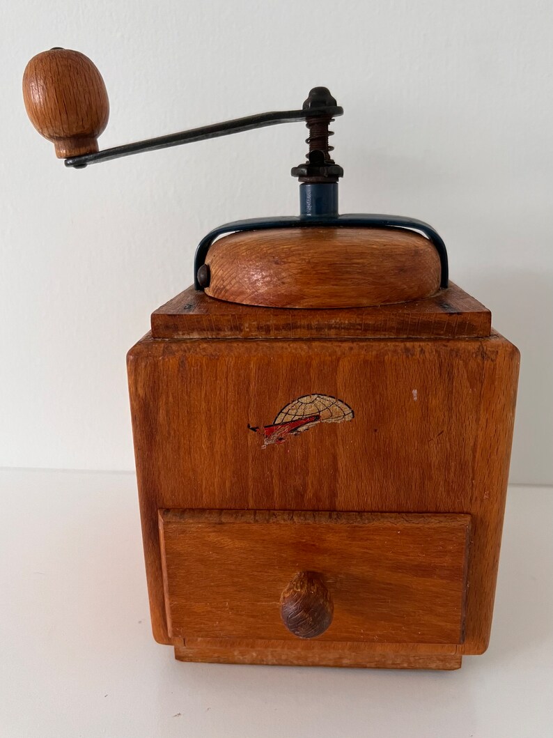 Puede incluir: Un molinillo de caf&eacute; de madera con cuerpo cuadrado, un peque&ntilde;o caj&oacute;n extra&iacute;ble con pomo redondo y una manivela de metal montada en la parte superior con un pomo de madera. El frente de la caja de madera muestra un peque&ntilde;o gr&aacute;fico desgastado de un globo.
