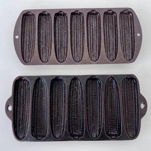 May include: Two cast iron corn stick pans. Each pan has a dark brown finish and features eight corn cob-shaped moulds. The pans have handles with holes for hanging. These pans are designed for baking corn bread in the shape of corn cobs.