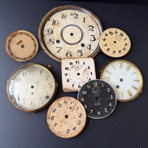 May include: A collection of eight vintage clock faces and dials in various shapes and sizes. The assortment includes round and square metal pieces featuring black Arabic and Roman numerals, some with aged patina, visible center holes, and brand text.