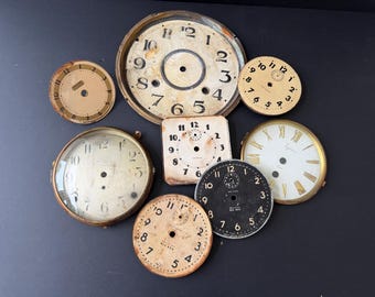 May include: A collection of eight vintage clock faces and dials in various shapes and sizes. The assortment includes round and square metal pieces featuring black Arabic and Roman numerals, some with aged patina, visible center holes, and brand text.