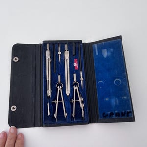 May include: A black leather case with a blue velvet interior holds a set of silver compass tools. The case has a silver snap closure and the words "GRAU" are printed on the front.