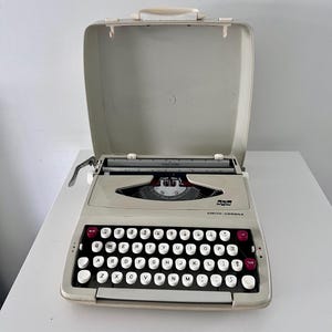 May include: A vintage Smith-Corona typewriter in white with a black keyboard and red keys. The typewriter is in a closed carrying case.