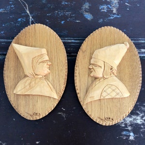 May include: Two wooden carvings of people's heads, each with a hat and a carved face. The carvings are on oval-shaped wooden plaques with a decorative border. The plaques are signed "Armin".