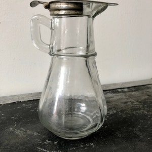 May include: A clear glass pitcher with a silver lid and a handle. The pitcher has a round body and a spout. It is sitting on a dark surface.