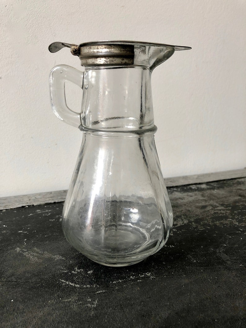 May include: A clear glass pitcher with a silver lid and a handle. The pitcher has a round body and a spout. It is sitting on a dark surface.