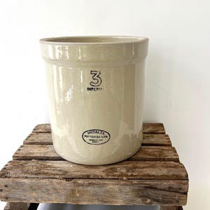 May include: A beige stoneware crock with a cylindrical shape, marked with a black "3" and "IMPERIAL". The crock also features a black oval stamp from Medalta Potteries Ltd. It sits on a rustic wooden crate, suggesting a vintage or farmhouse aesthetic.
