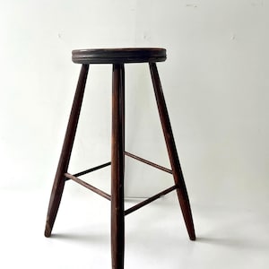 May include: A dark brown wooden stool with a round seat and three angled legs. The stool has a simple design and appears to be vintage or antique. The legs are connected by a horizontal support.