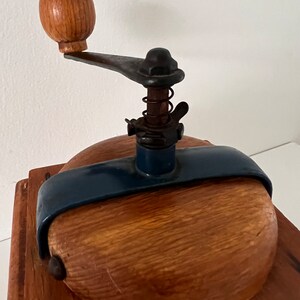 Puede incluir: Un molinillo de caf&eacute; manual con base cuadrada de madera, tapa redondeada de madera y una abrazadera met&aacute;lica de color azul oscuro. El mecanismo incluye una varilla met&aacute;lica vertical con un resorte, una tuerca de mariposa y un pomo de madera.