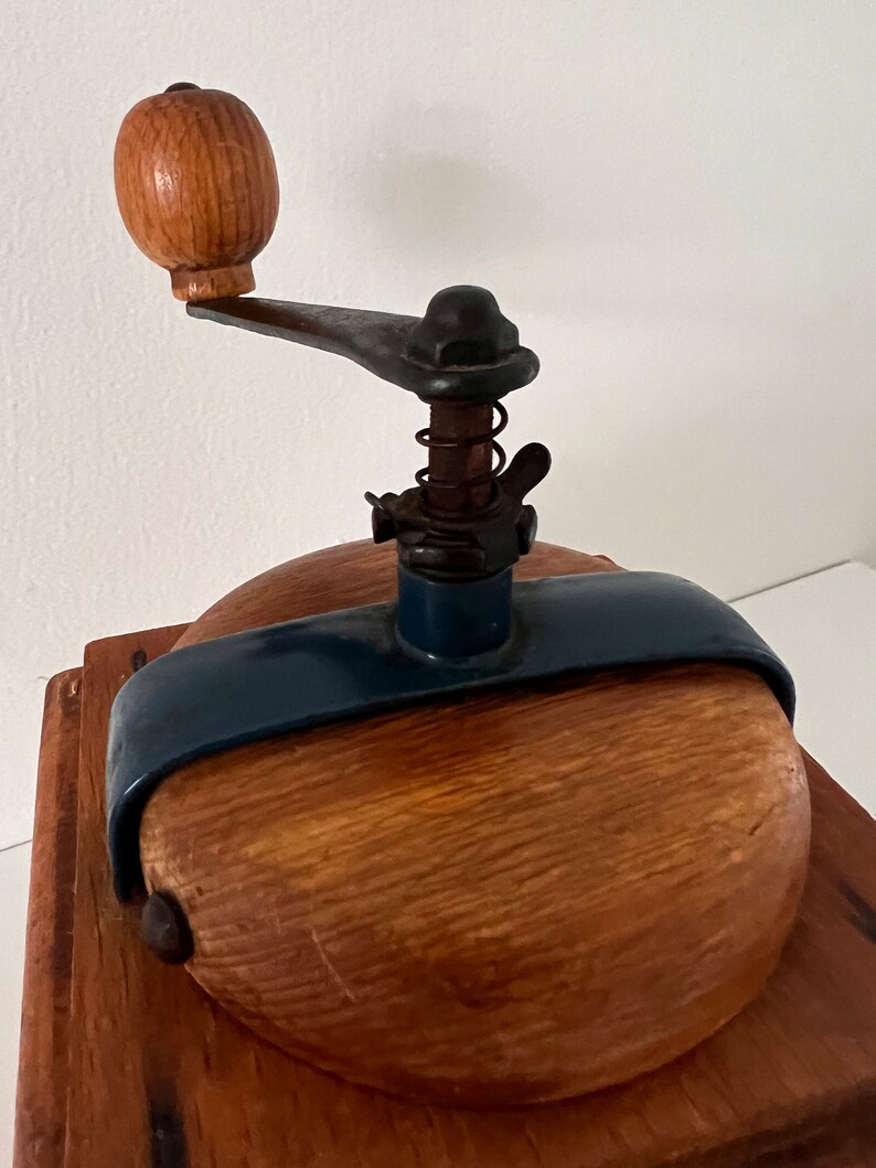 Puede incluir: Un molinillo de caf&eacute; manual con base cuadrada de madera, tapa redondeada de madera y una abrazadera met&aacute;lica de color azul oscuro. El mecanismo incluye una varilla met&aacute;lica vertical con un resorte, una tuerca de mariposa y un pomo de madera.