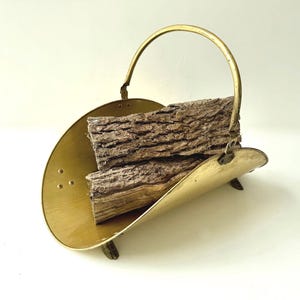Peut inclure: Un porte-bûches en laiton poli avec un design incurvé à extrémités ouvertes et une poignée arquée unique. Le support repose sur trois petits pieds et contient deux morceaux de bois de chauffage brun, rugueux et texturé, empilés à l'intérieur.