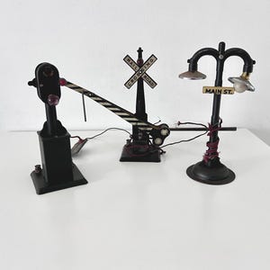 May include: A vintage toy train crossing set with a black and white striped gate, a red and black signal light, and a black street lamp with a sign that reads "MAIN ST.".