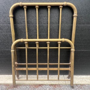 May include: A vintage brass bed frame with a headboard and footboard. The frame has vertical bars and rounded top rails. The bed frame is made of brass and has a warm, golden color. The bed frame is designed for a standard size mattress.